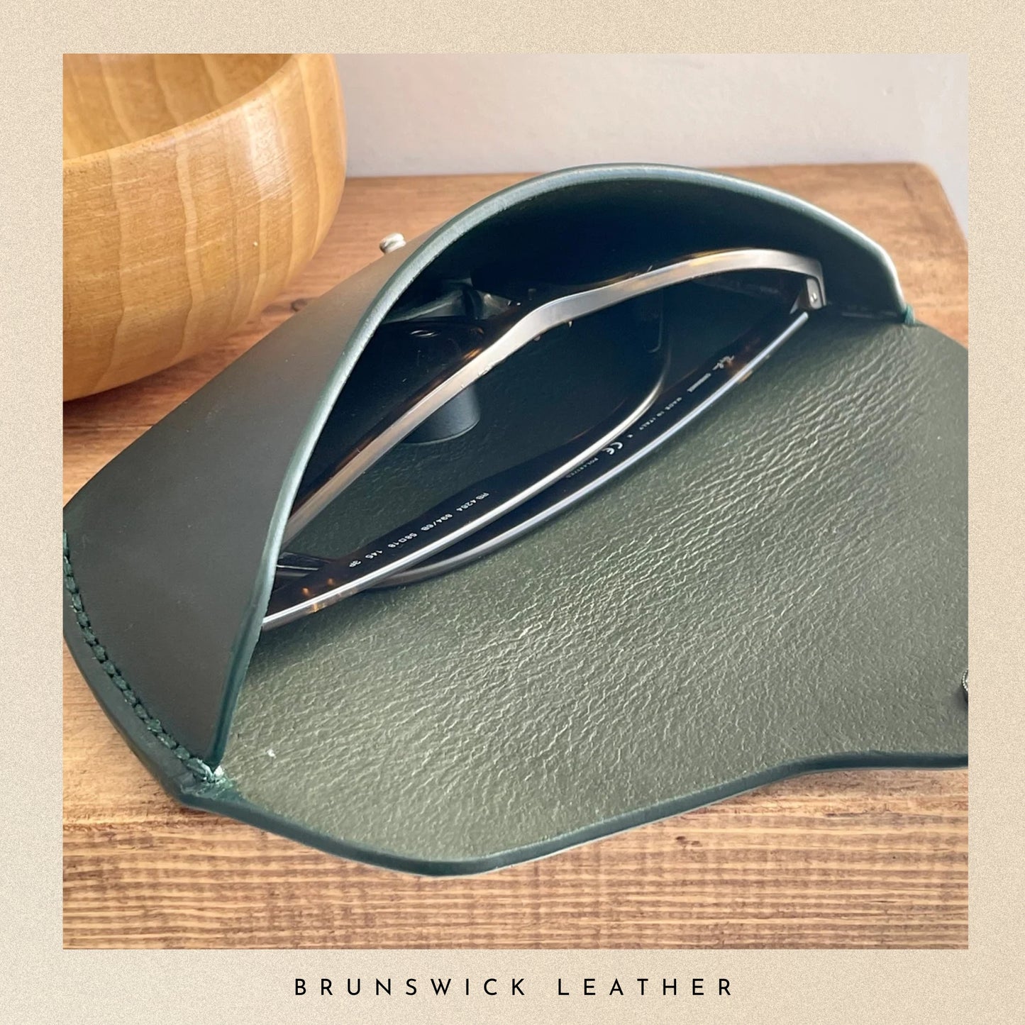 Green leather sunglasses case with sunglasses on a wooden surface, featuring 'Brunswick Leather' branding.