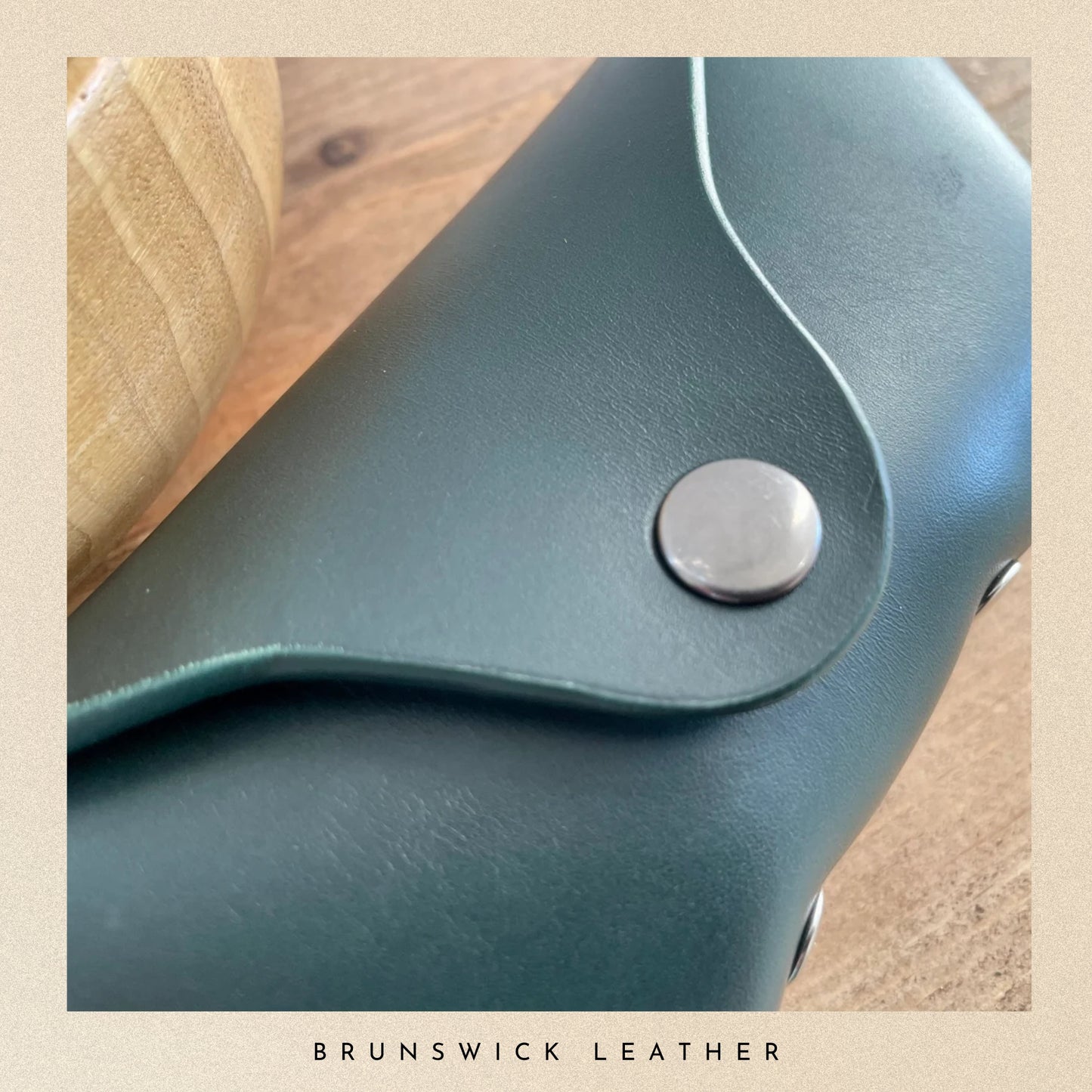 Close-up of teal leather product with a silver button on a wooden surface, branded 'Brunswick Leather'.