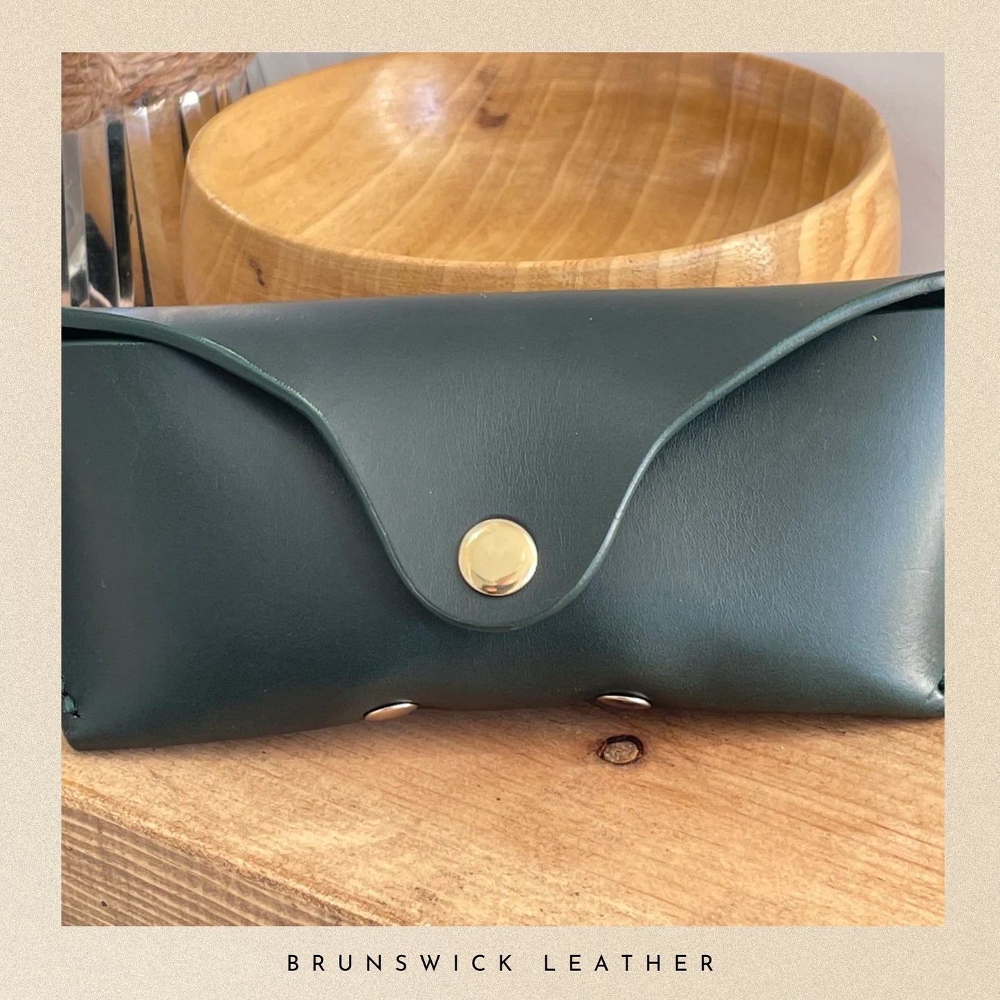 Green leather clutch with gold clasp on a wooden surface, branded 'Brunswick Leather'.