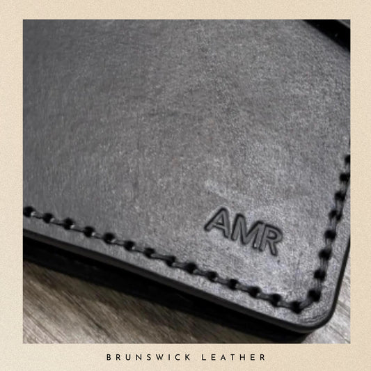Gray leather wallet with 'AMR' branding on a wooden surface, branded 'Brunswick Leather'.