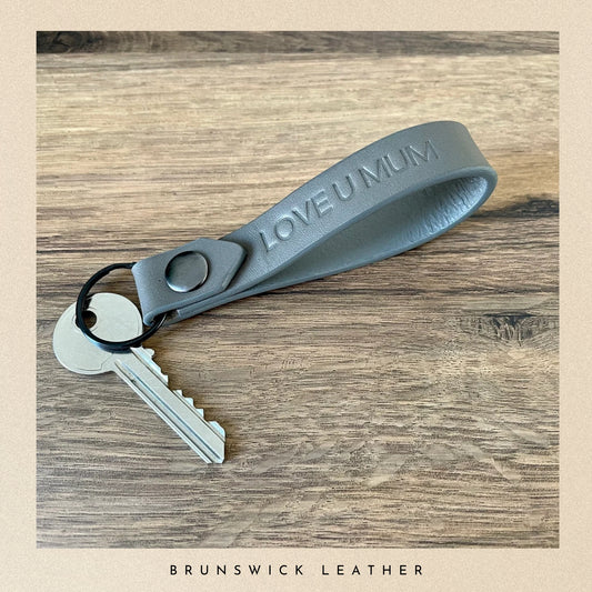 Grey leather keychain with 'LOVE U MUM' engraving on a wooden surface, branded 'Brunswick Leather'.
