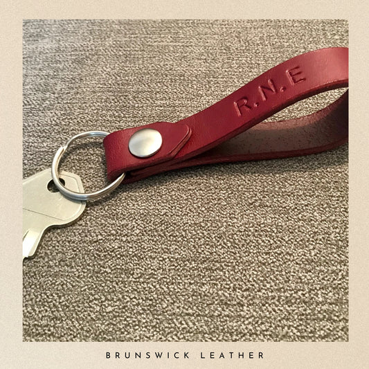 Burgundy leather keychain with 'R.N.E.' engraving on a textured surface, branded 'Brunswick Leather'.