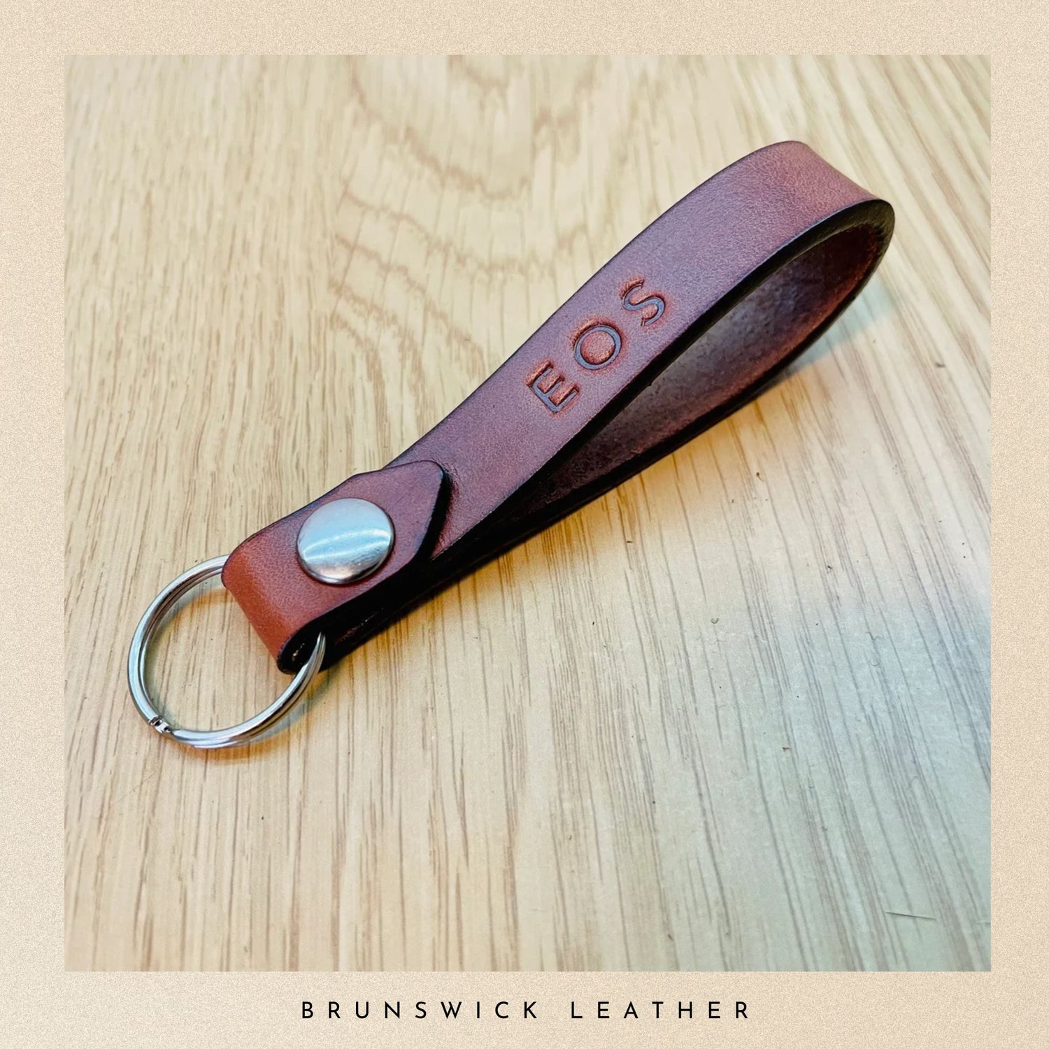 Brown leather keychain with 'EOS' engraving on a wooden surface, branded 'Brunswick Leather'.
