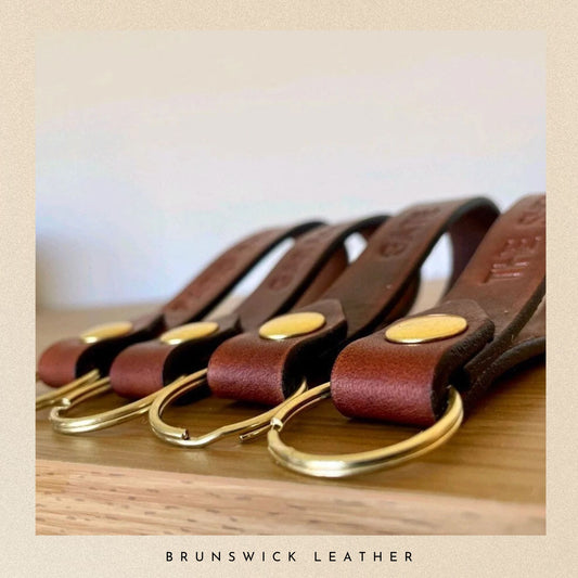 Set of leather keychains with gold clasps on a wooden surface, branded 'Brunswick Leather'.