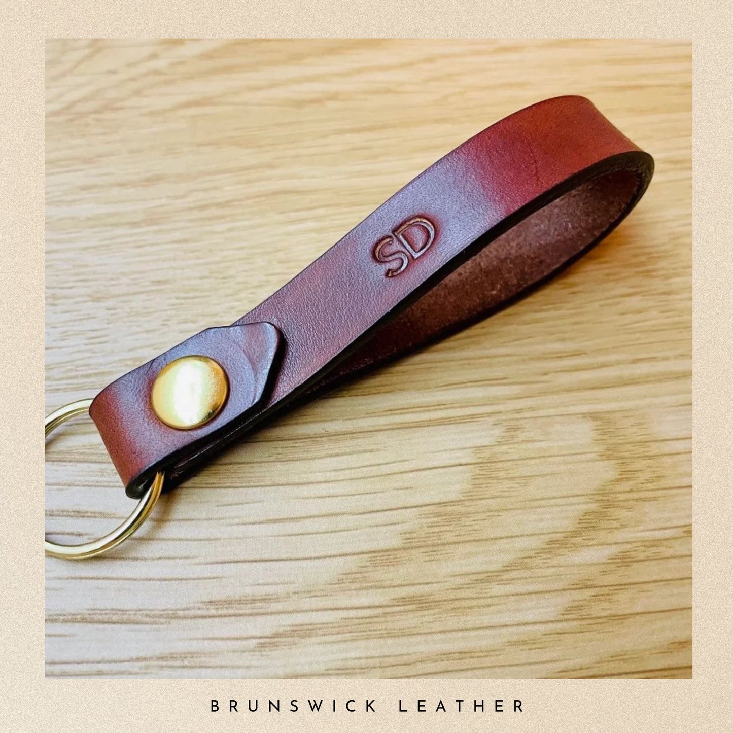 Brown leather keychain with gold accent on a wooden surface, branded 'Brunswick Leather'.