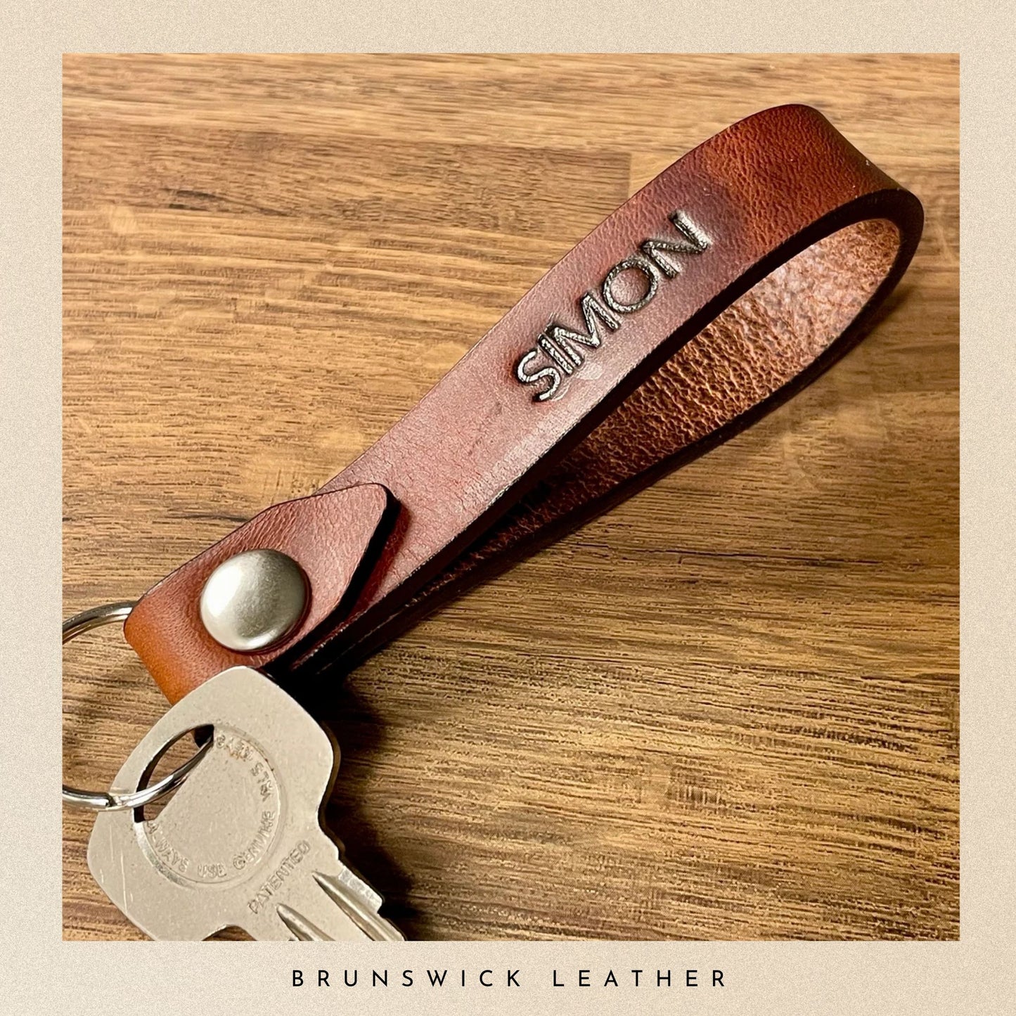 Brown leather keychain with 'SIMON' engraving on a wooden surface, branded 'Brunswick Leather'.