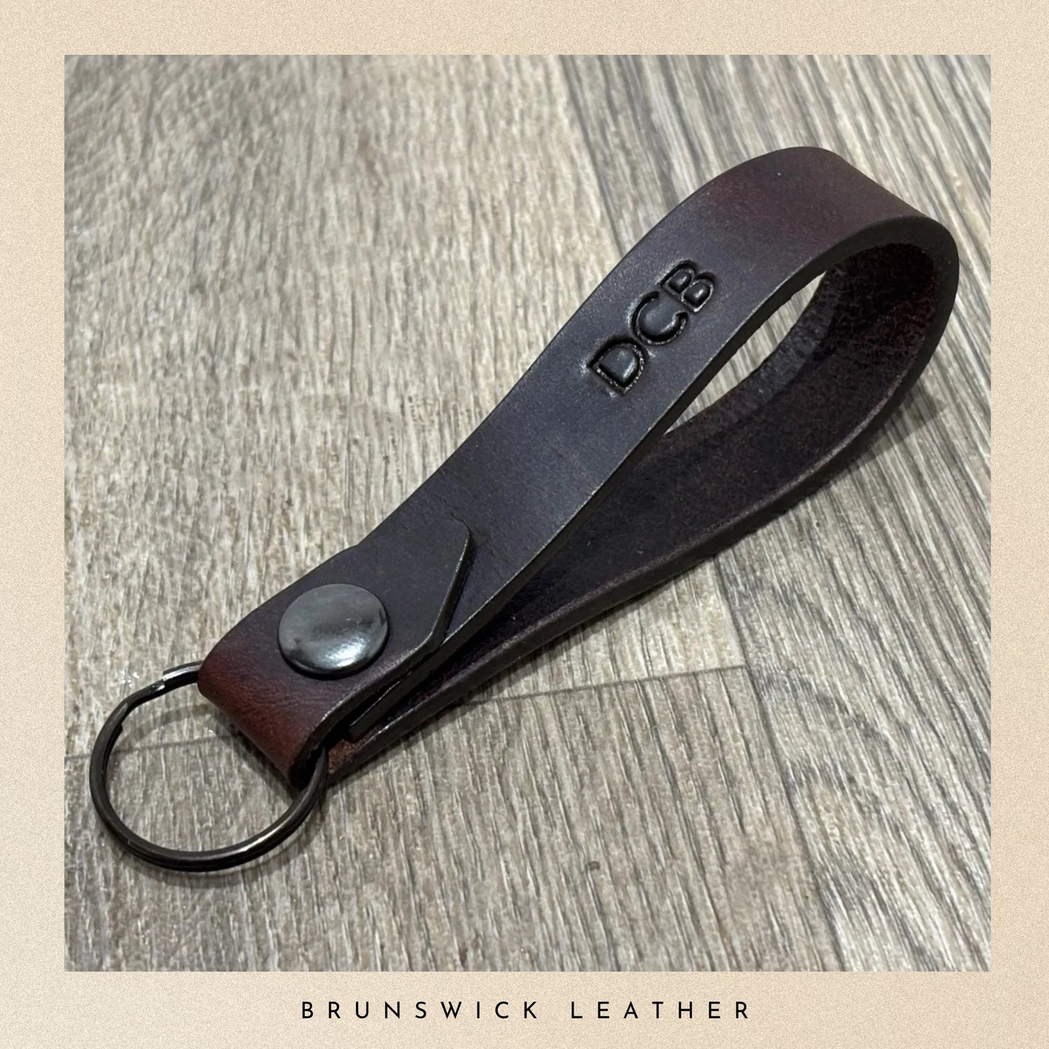 Brown leather keychain with 'DCB' branding on a wooden surface, branded 'Brunswick Leather'.