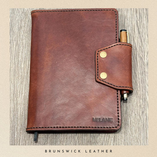Brown leather notebook with pen holder on a wooden surface, branded 'Melane'.