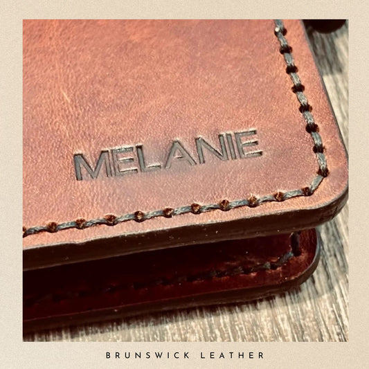 Brown leather wallet with 'Melanie' engraving on a wooden surface, branded 'Brunswick Leather'.