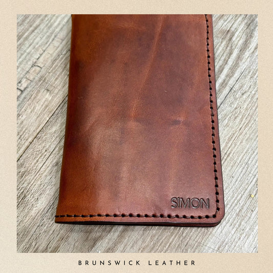 Brown leather wallet with 'Simon' branding on a wooden surface, featuring the brand 'Brunswick Leather'.
