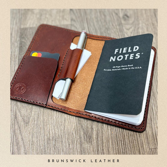 Brown leather wallet with a pen, cardholder, and Field Notes notebook on a wooden surface.