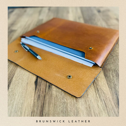 Brown leather notebook cover with pens on a wooden surface, branded 'Brunswick Leather'.