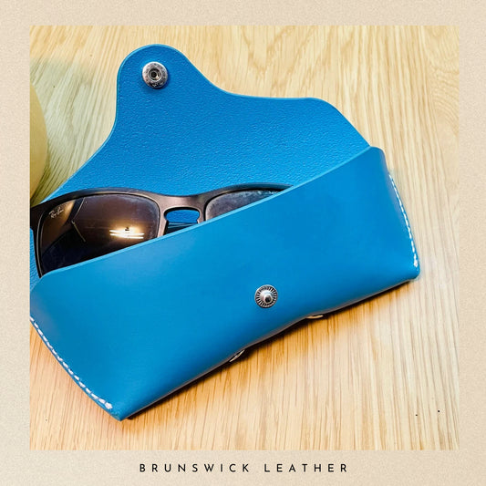 Blue leather sunglasses case with sunglasses inside on a wooden surface, branded 'Brunswick Leather'.