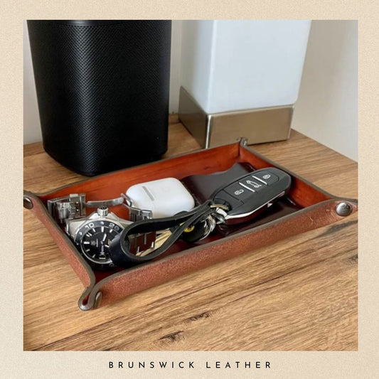 Brown leather tray with watch, keys, and phone on a wooden surface, branded 'Brunswick Leather'.
