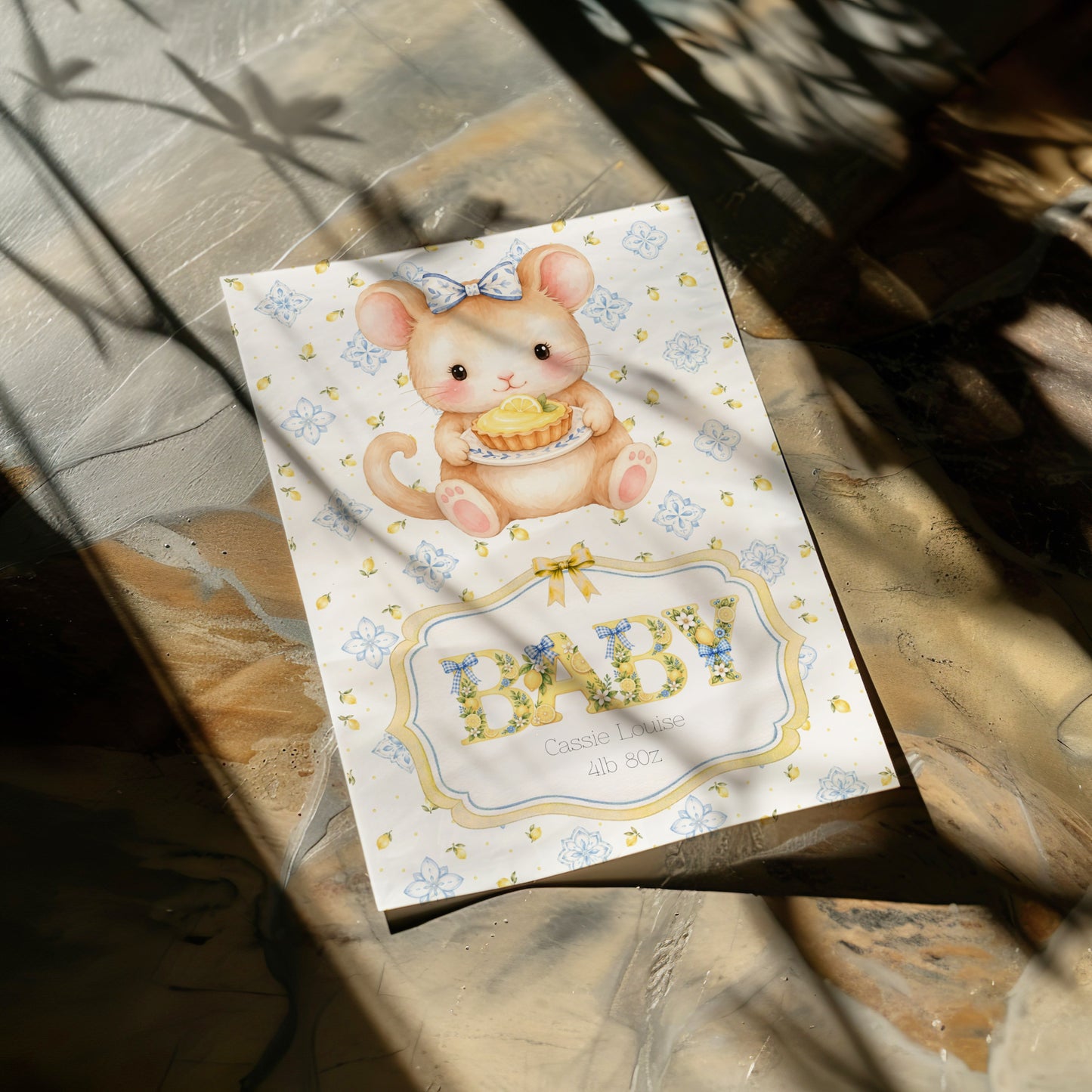 A baby card featuring a cute mouse holding a yellow basket, with the word 'BABY' in floral letters, photographed on a marble surface with dappled sunlight and shadows.