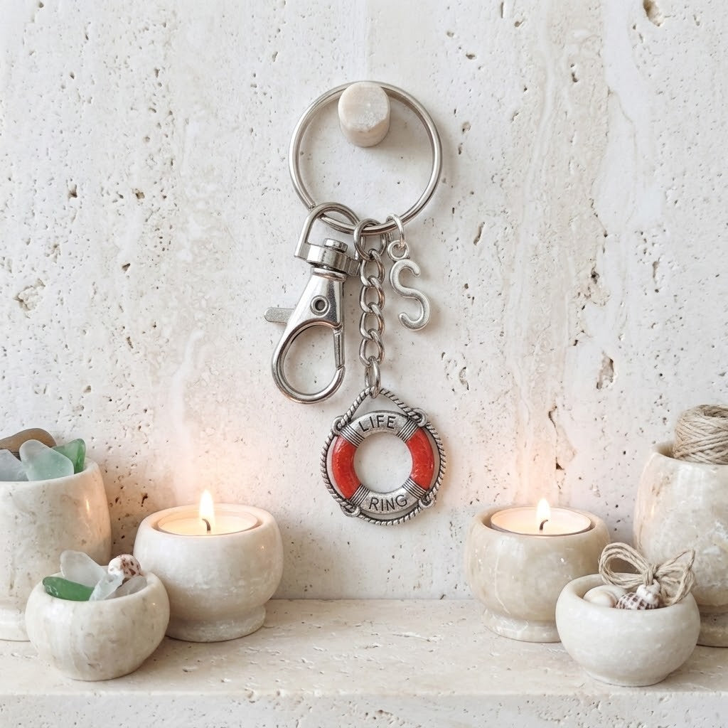 A silver-toned metal keychain with a lobster clasp, key ring, and a life preserver charm featuring red and white lettering, displayed on a textured cream wall above a shelf with lit candles and small decorative bowls.