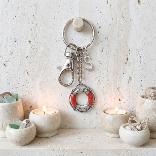 A silver-toned metal keychain with a lobster clasp, key ring, and a life preserver charm featuring red and white lettering, displayed on a textured cream wall above a shelf with lit candles and small decorative bowls.
