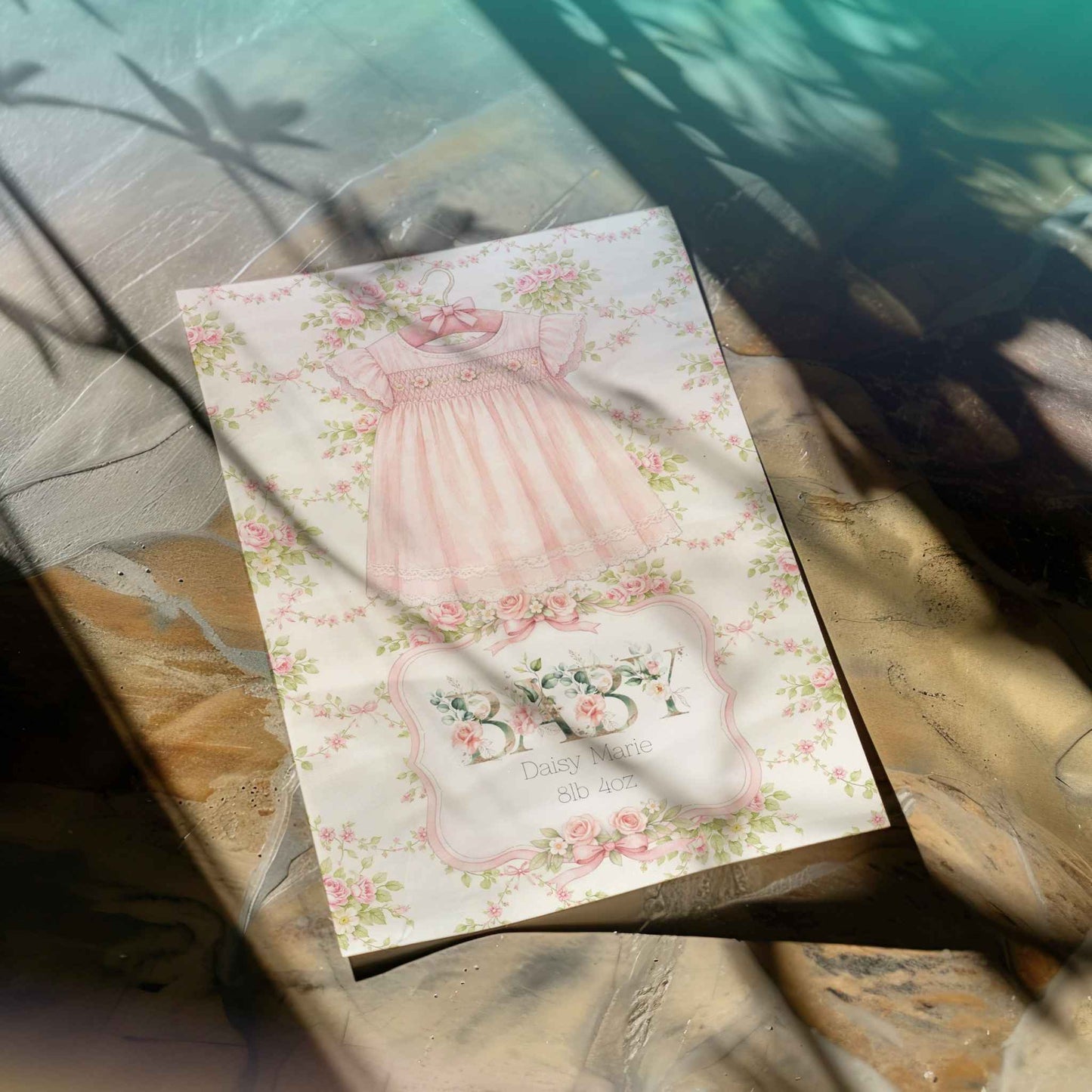 A decorative printed card or art print featuring a pastel pink dress illustration and floral border, placed on a stone surface with dappled sunlight and shadows.