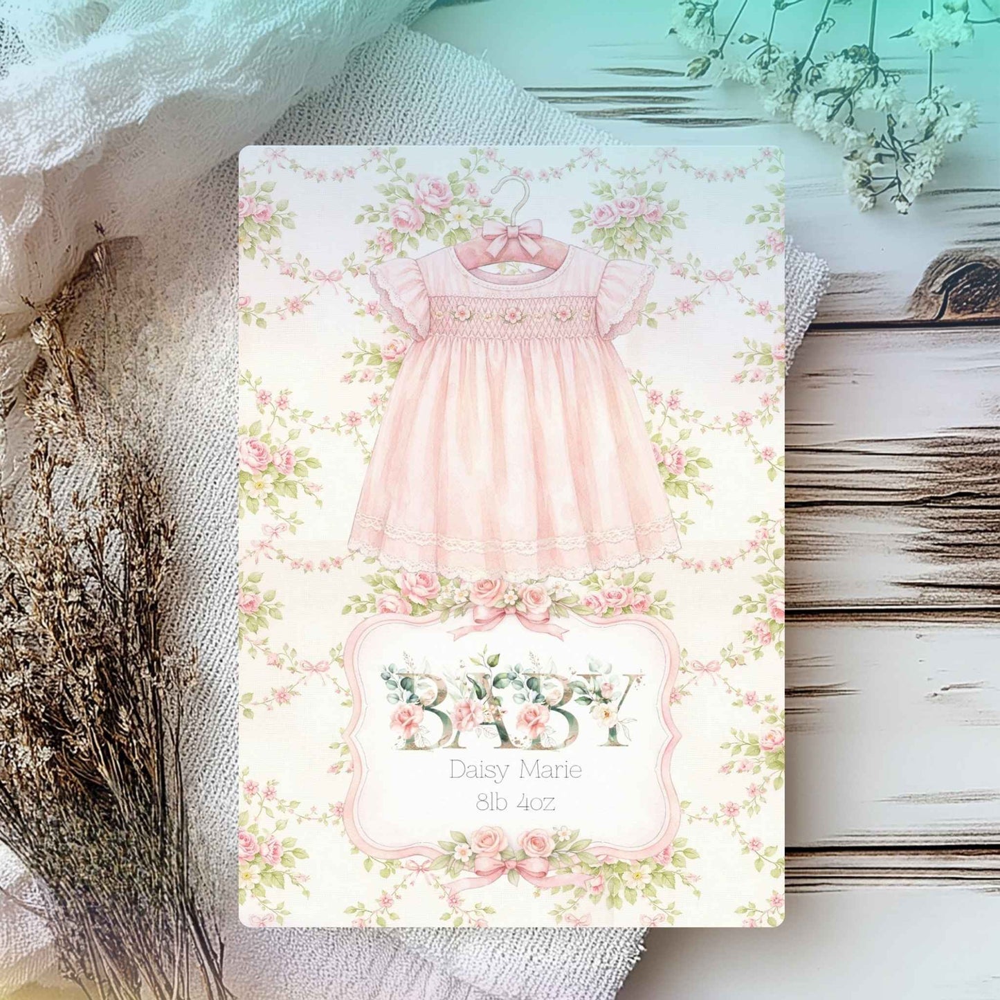 A baby shower card featuring a watercolor illustration of a pink baby dress on a floral background, styled on a rustic wooden surface with dried flowers and fabric.