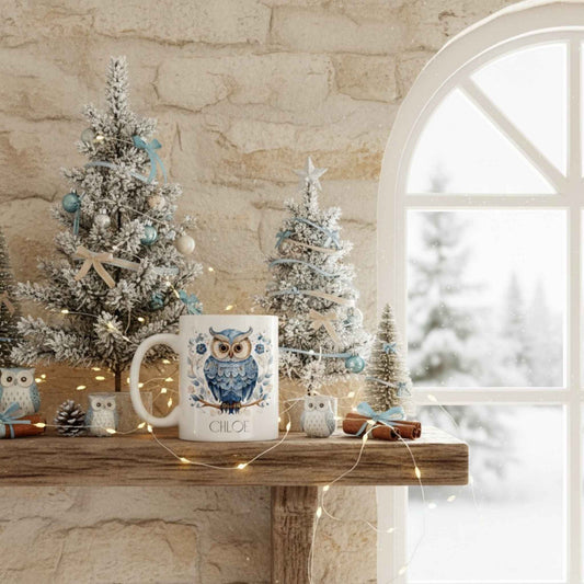 Decorative scene with a mug featuring an owl design on a wooden surface with small Christmas trees and lights.