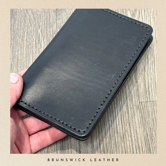 Black leather wallet held in a hand on a wooden surface with 'Brunswick Leather' branding.