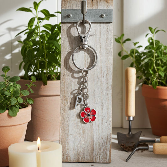 A silver metal keychain with a red flower charm and a letter 'P' charm hanging from a rustic white wooden peg holder, styled with potted plants, a lit candle, and gardening tools.