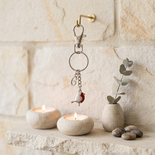 A silver keychain with a small red robin bird charm hangs from a brass hook mounted on a light stone wall. Below, a stone ledge holds two lit tea lights, a small vase with eucalyptus, and a few smooth stones.