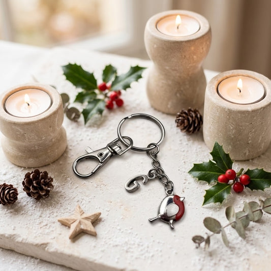 A silver-toned metal keychain with a small bird charm and a red accent, displayed on a rustic white surface surrounded by lit candles, pinecones, holly leaves, and berries.