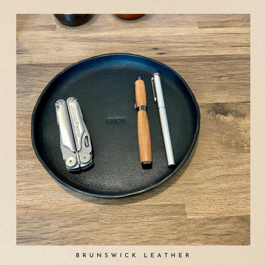 Black leather round personalised valet tray with a pen and a tool on a wooden surface, branded 'Brunswick Leather'. 