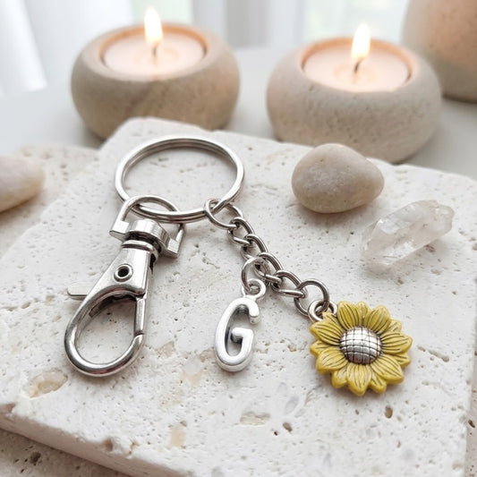 A silver metal keychain with a lobster clasp, a hanging letter 'G' charm, and a yellow sunflower charm, displayed on a textured white stone with lit candles in the background.