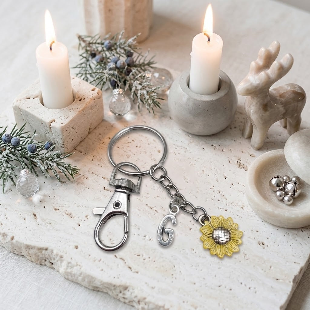 A silver-toned keychain with a lobster clasp, a letter 'G' charm, and a yellow sunflower charm, styled on a stone surface with lit candles, pine branches, a deer figurine, and small silver beads.