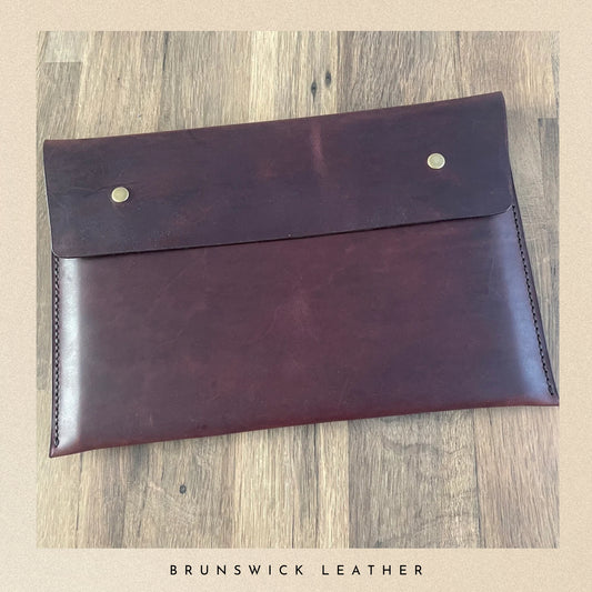 Brown leather envelope with metal studs on a wooden surface, branded 'Brunswick Leather'.