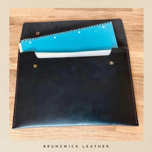 Black leather pouch with a blue notebook inside on a wooden surface, branded 'Brunswick Leather'.