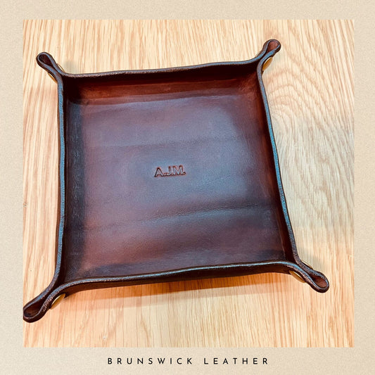 Brown leather tray with 'AJM' branding on a wooden surface, branded 'Brunswick Leather'.