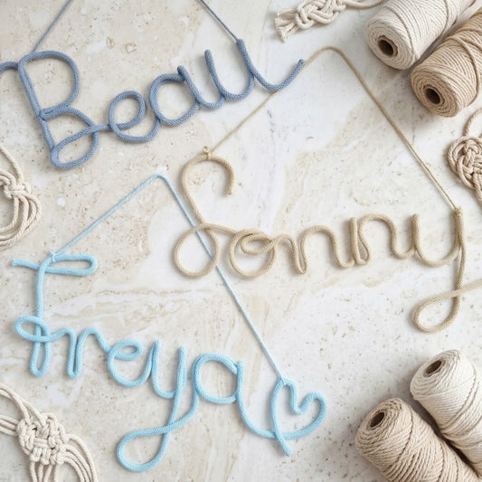 Three cursive hanging name banners in light blue and natural beige yarn, arranged on a light stone surface with spools of twine and macrame decor around them.