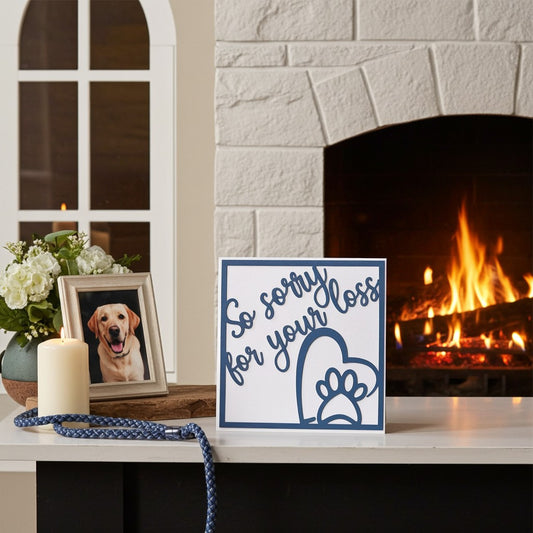 A square white memorial card with blue text reading 'So sorry for your loss' and a blue paw print outline, displayed on a fireplace mantel beside a candle, flowers, a framed dog photo, and a blue rope. A real indoor home environment with a burning fireplace is visible