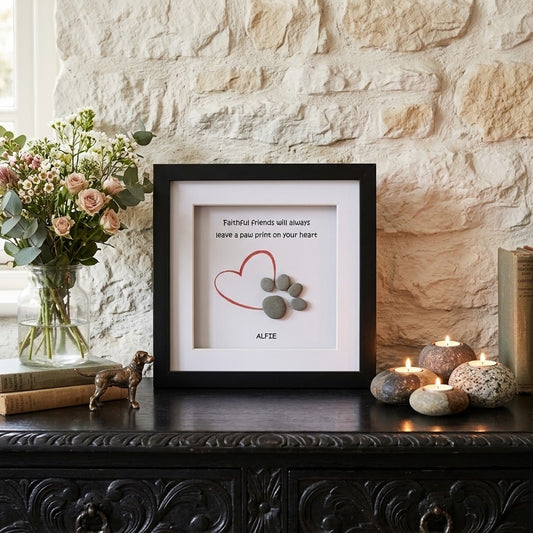 A framed paw print pebble art wall art piece displayed on a dark carved wooden console table against a textured stone wall. The black frame contains a white background with a red heart outline, a paw print made of stones, and the text 'Famous friends will share leave a paw print on your heart' above the name 'ALFIE'.