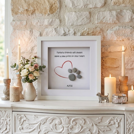 A framed pet memorial pebble art displayed on a white carved console table against a stone wall. The white frame contains a red heart outline, grey paw prints, the quote 'Faithful Friends will always Leave a paw print on your heart', and the name 'ALFIE'.