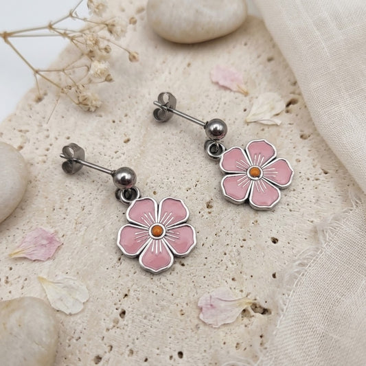 A pair of silver-tone dangle earrings with pink flower charms and small orange centers, displayed on a textured beige stone surface with scattered petals and fabric.