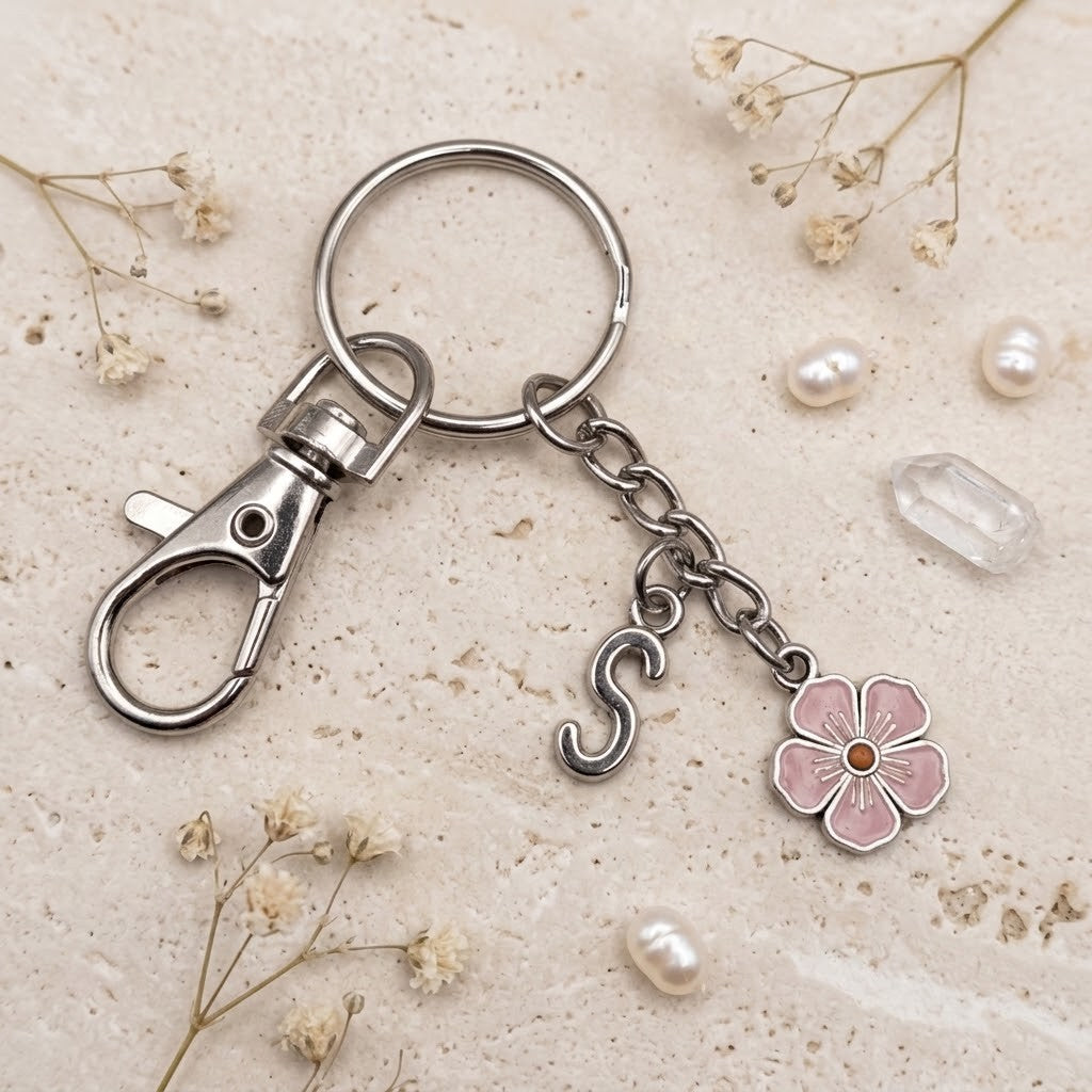 A silver-tone metal keychain with a lobster clasp, split ring, chain, a hanging letter 'S' charm, and a pink flower charm, styled on a textured beige surface with dried flowers and pearls.