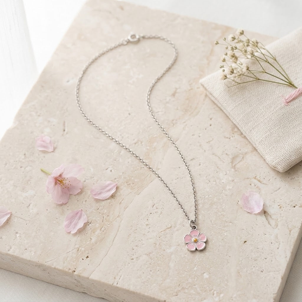 A delicate silver chain necklace with a small pink flower pendant, styled on a beige stone slab with scattered pink petals and a small bunch of baby's breath.