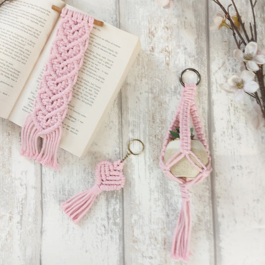 A set of handmade macrame crafts including a keyring, bookmark, and mini plant hanger, primarily in pink colour, displayed on a wooden surface with a background of flowers and books.