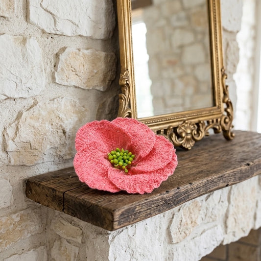 A pink crocheted flower with a green centre sits on a rustic wooden mantel against a white stone wall, with an ornate gold mirror reflected behind it.