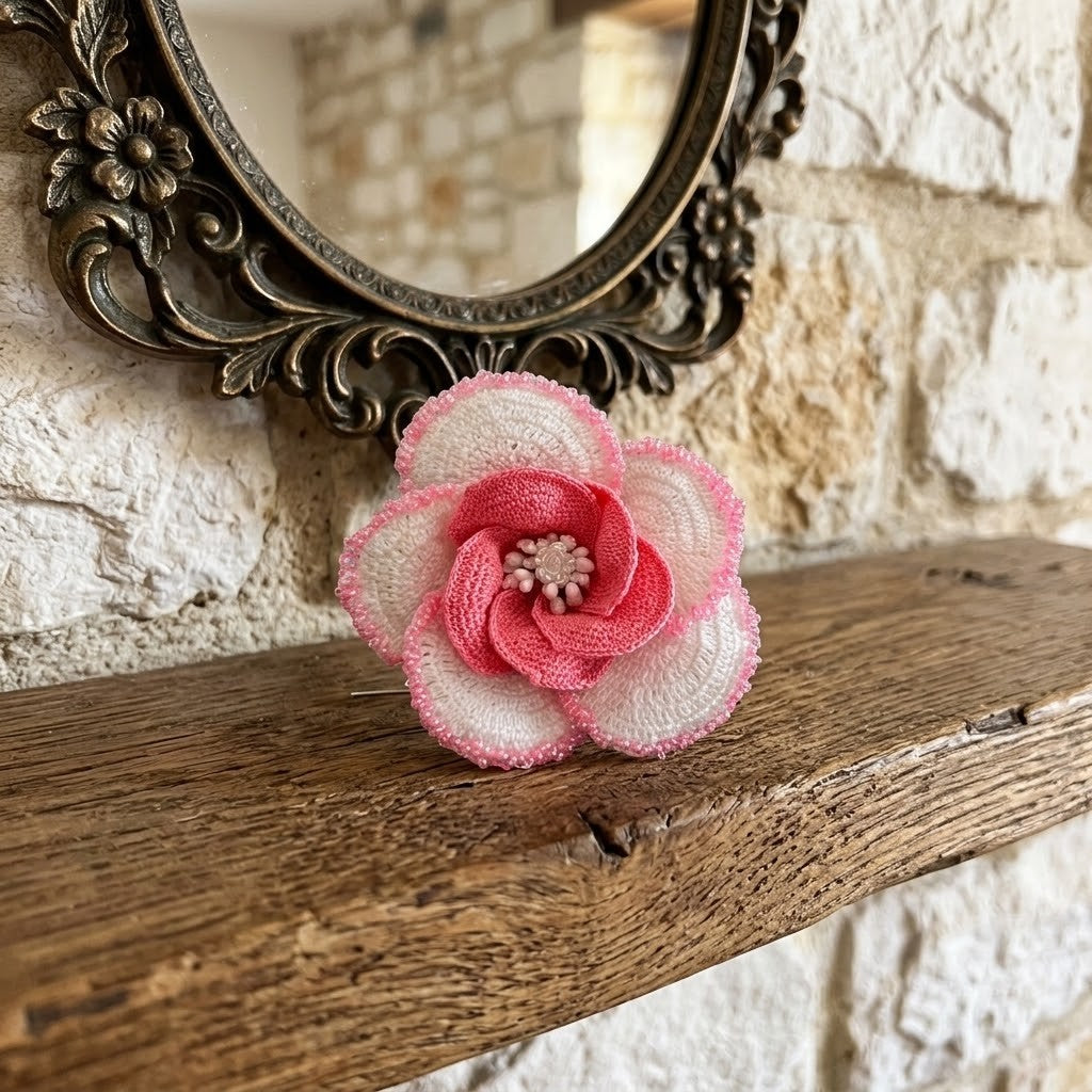 A handmade fabric flower brooch with pink and white petals sits on a rustic wooden mantelpiece in front of a stone wall and an ornate mirror.