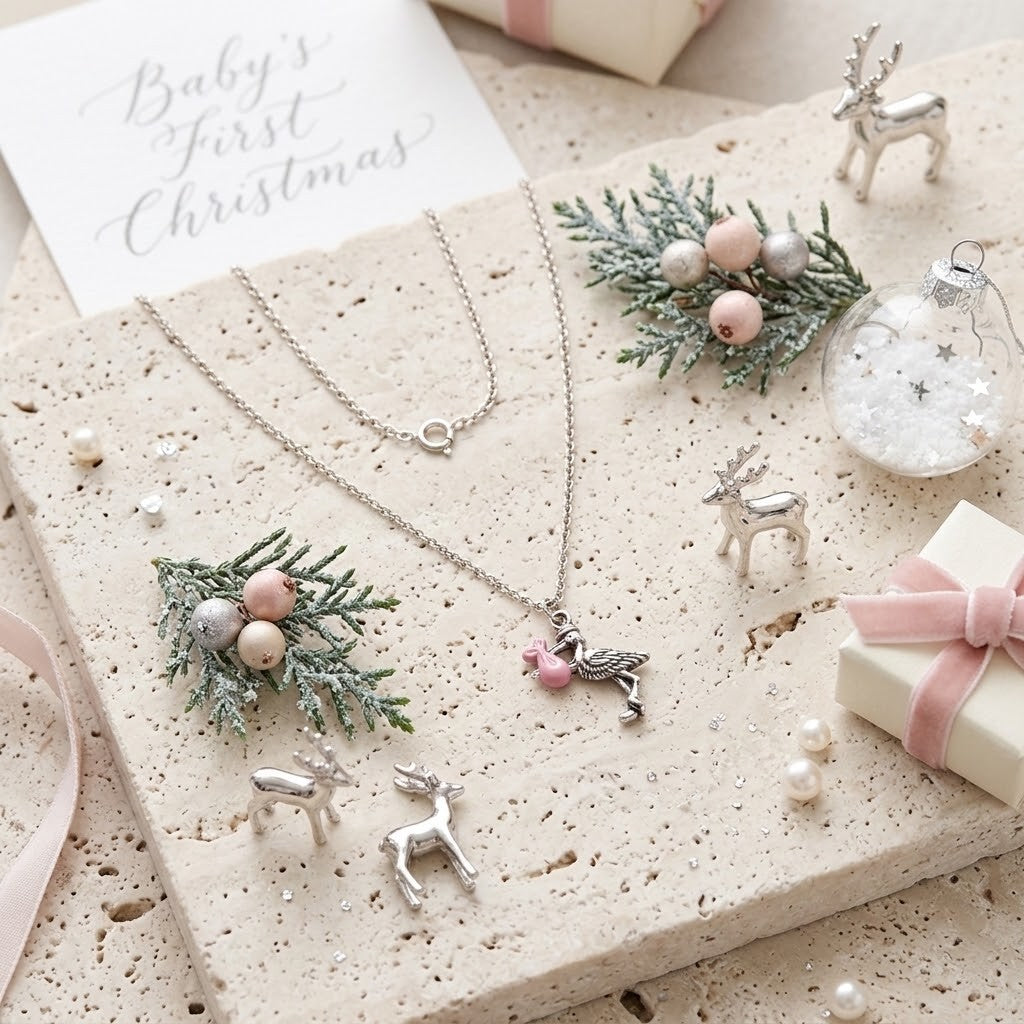 Silver baby first Christmas necklace set with pink stork pendants, styled on a textured stone slab with festive ornaments, a gift box, and a card reading 'Baby's First Christmas'.