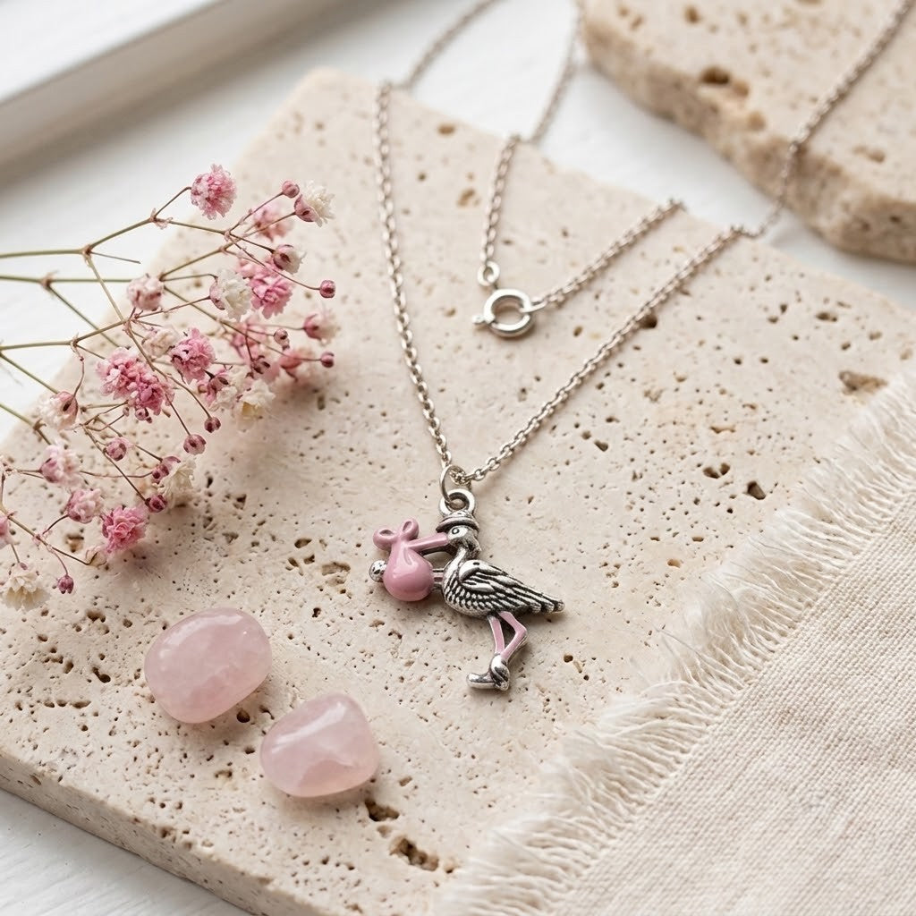 A silver-toned necklace with a pink stork pendant featuring pink enamel details, styled on a textured beige stone slab with pink stones and dried pink flowers.