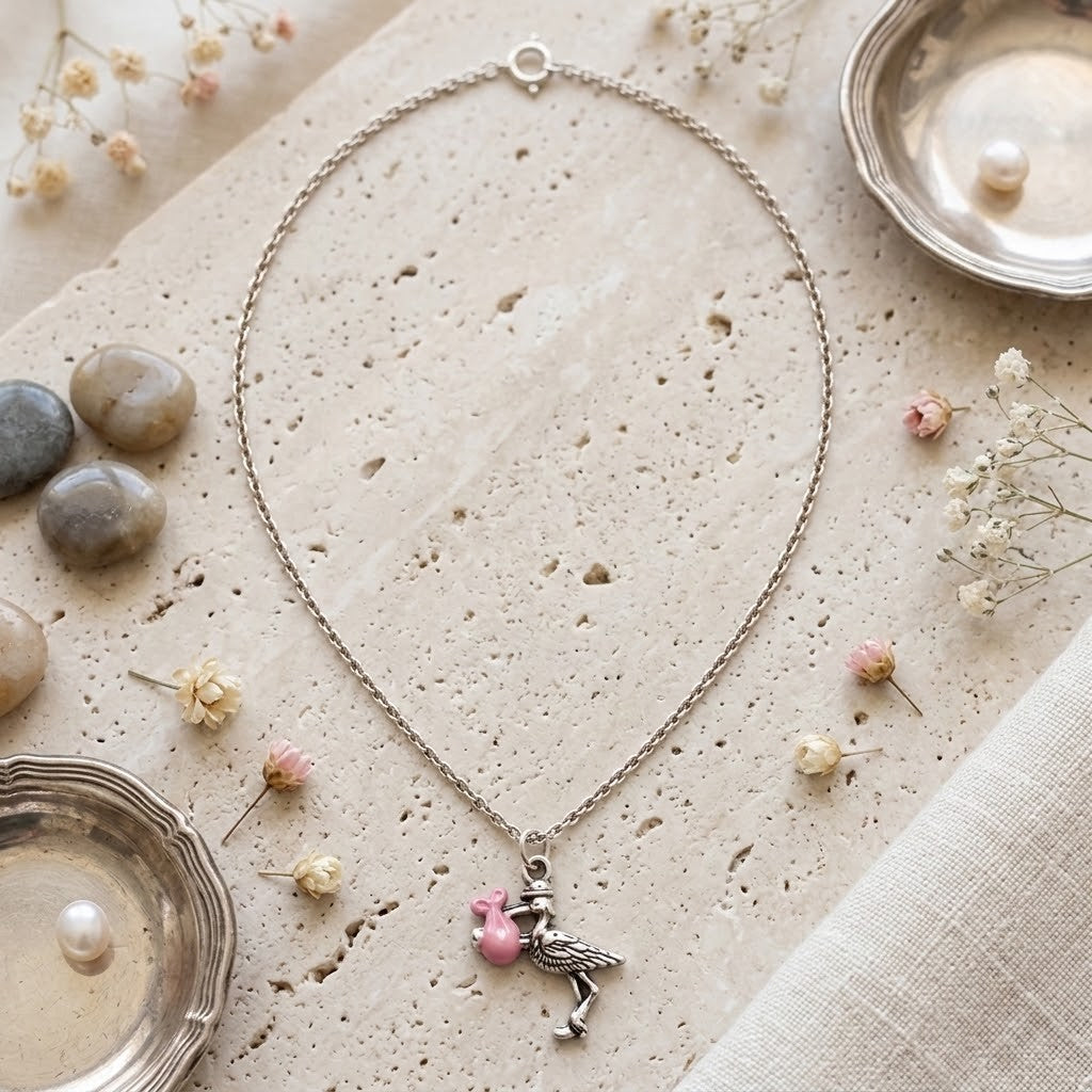 A silver chain necklace with a stork charm holding a pink heart pendant, styled on a textured beige stone surface with dried flowers, small stones, and silver dish props.