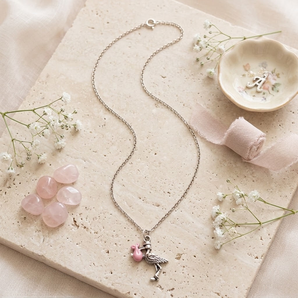 A silver-toned chain necklace with a stork pendant, styled on a beige stone slab with pink rose quartz stones, white baby's breath flowers, a rolled pale pink ribbon, and a small floral dish.
