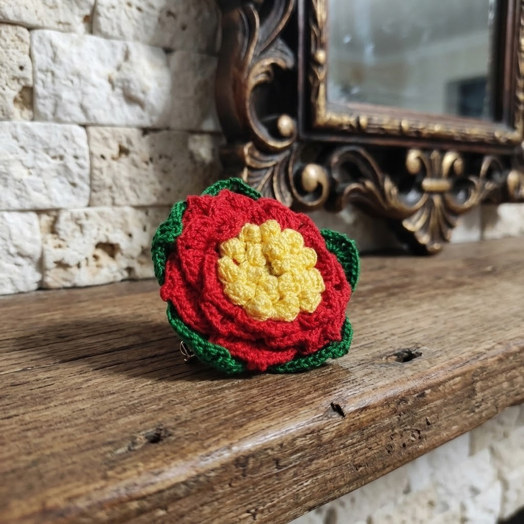 A crocheted flower hair clip with a red outer layer, yellow textured center, and green inner layer, resting on a rustic wooden mantelpiece in front of a stone wall and an ornate mirror.