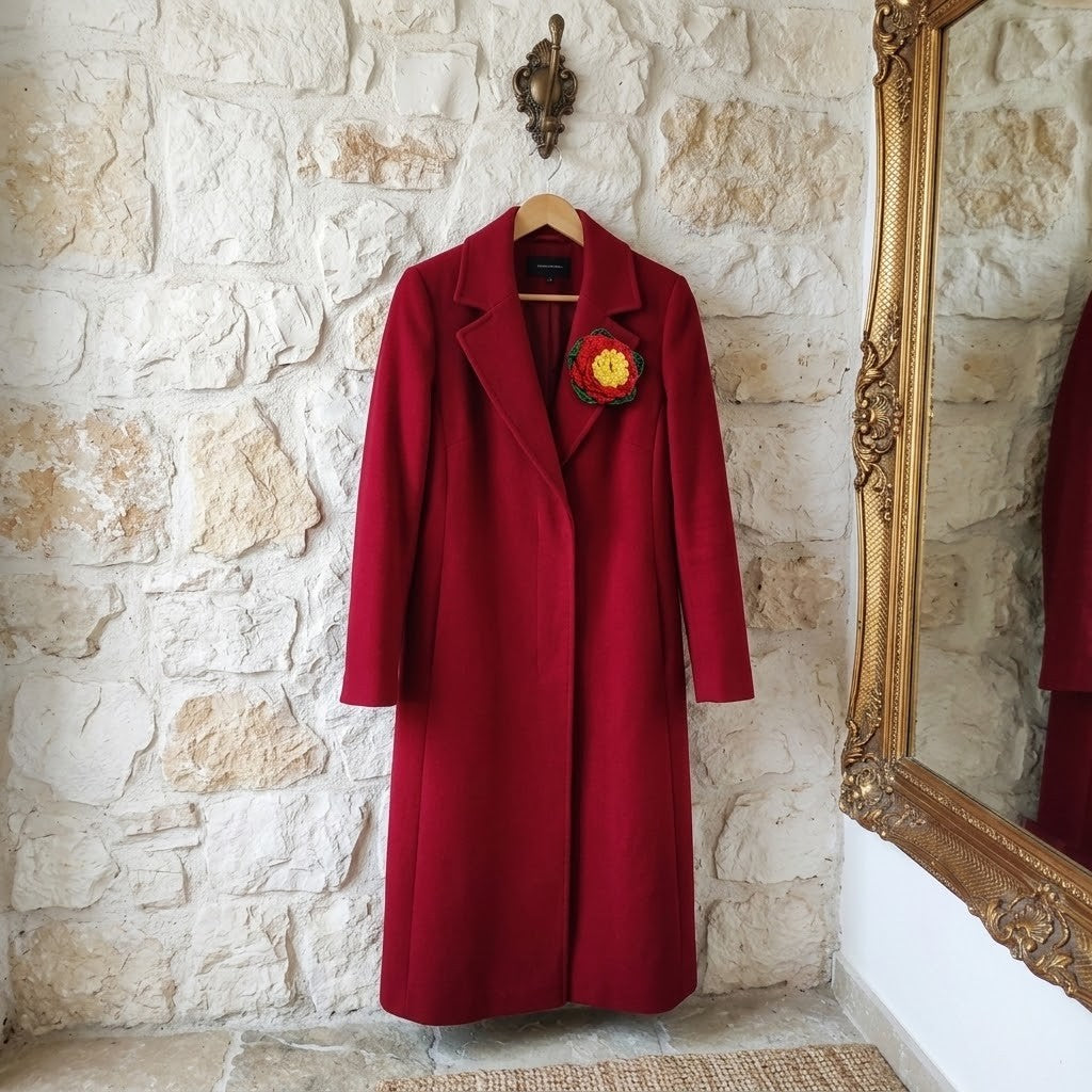 A long, deep red wool coat hangs on a wooden hanger against a rustic white stone wall, featuring a red and yellow embroidered floral lapel pin. A large ornate gold mirror is partially visible on the right side.
